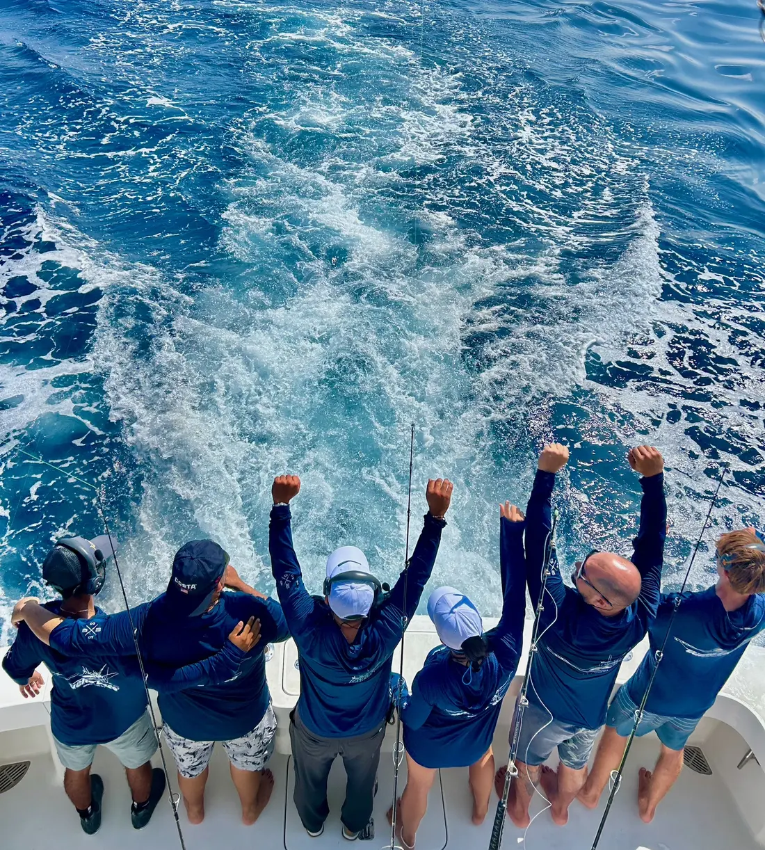 A crew of anglers on a Vuelto Loco charter boat raising their fists in celebration with the boat's wake visible behind them.
