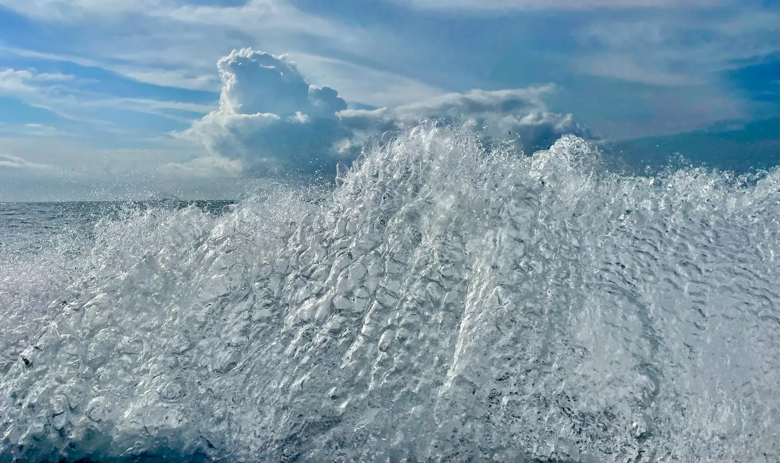 Close view of white spray and foam thrown up beside a moving boat, with a tall cumulus cloud over an open ocean horizon in the background.